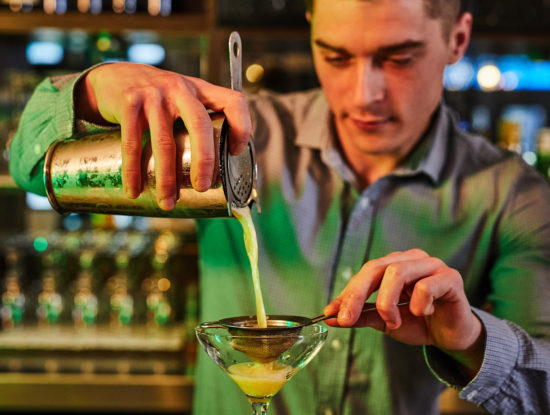 Bartender is pouring cocktail into a martini glass.
