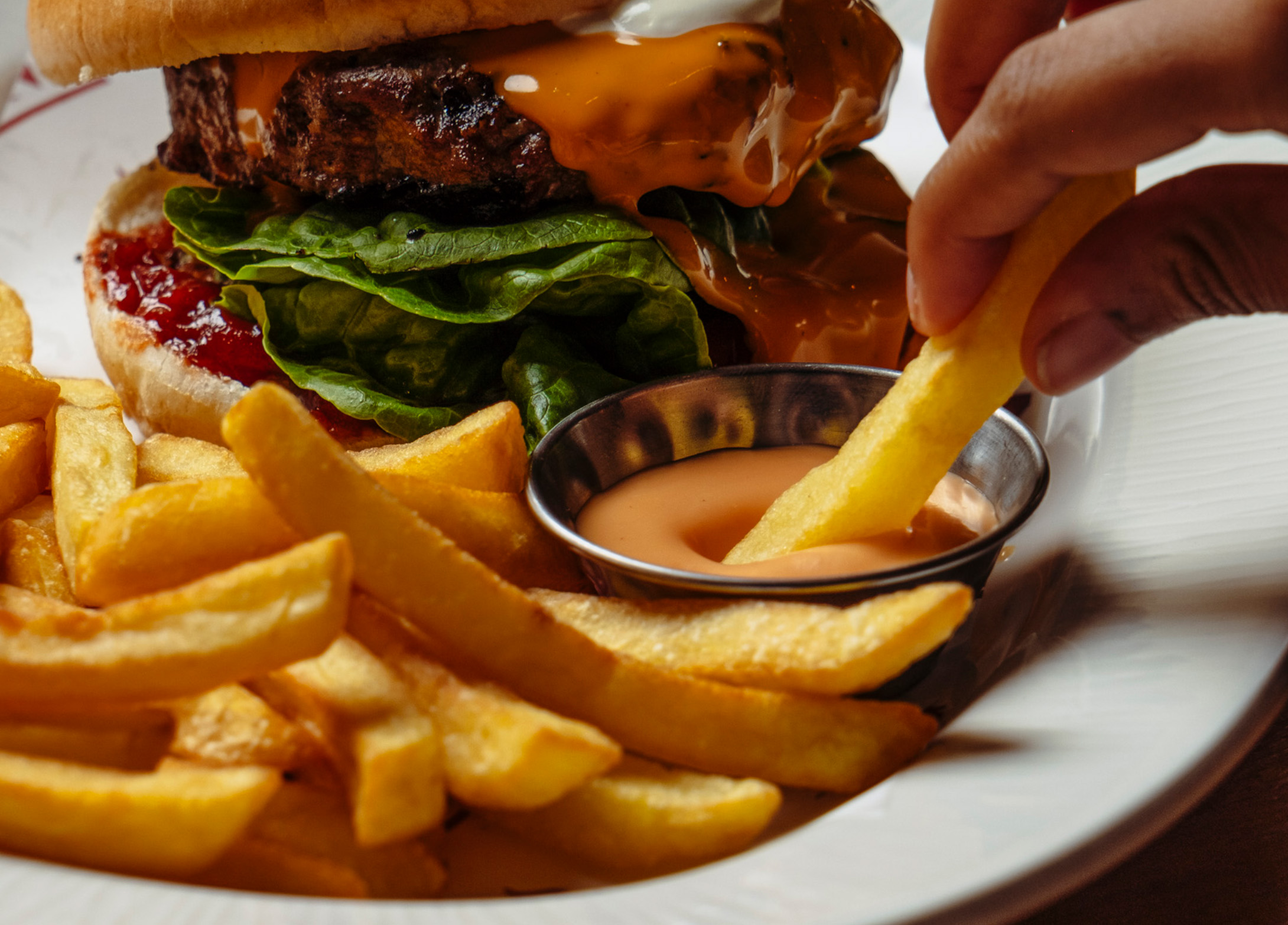 A person is dipping chips into a cheesy sauce with a delicious hamburger in the background.